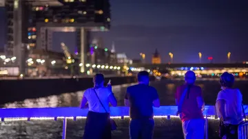 Menschen stehen am Wasser und betrachten die beleuchtete Stadt.