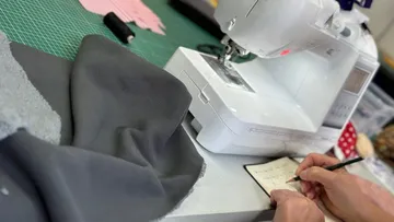 Nähmaschine auf Tisch, Person schreibt neben grauem Stoff.