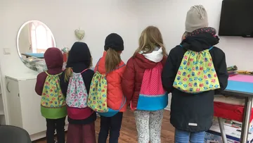 Five children stand back to back, wearing colorful backpacks.