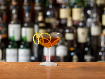 Cocktailglas mit orangefarbener Schleife, vor einer Bar.