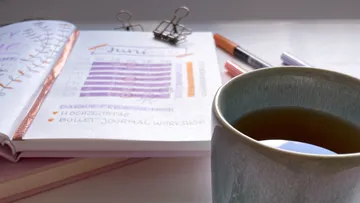 Tasse hält Tee, neben einem offenen Notizbuch und Stiften.