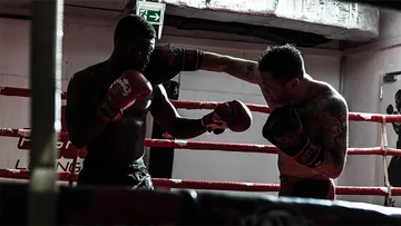 Two boxers fight in a ring with red ropes.