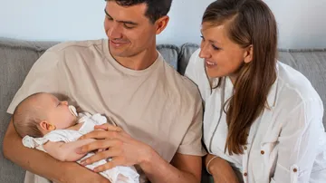 Parents hold sleeping baby, smile on the couch.