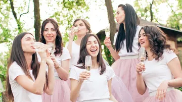 Six women drink sparkling wine and laugh outdoors.