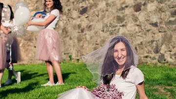 Woman with flowers sitting on grass smiling; other people are standing.