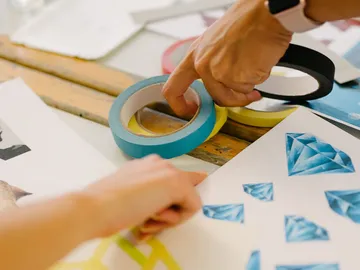 Hände greifen nach Malerband auf einem Tisch mit Papier.