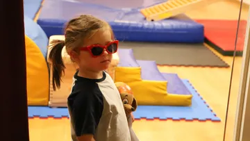 A child wears sunglasses and keeps a toy in a play room.