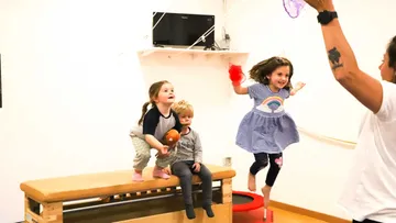 Zwei Kinder springen fröhlich von einer Bank in einem Raum.
