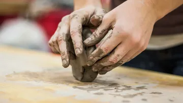 Hands form clay on a workbench-like surface.