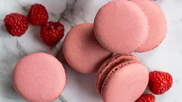 Rosa Macarons liegen auf Marmor mit Himbeeren daneben.