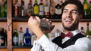 A bartender shakes a cocktail in front of a wall of shelves.