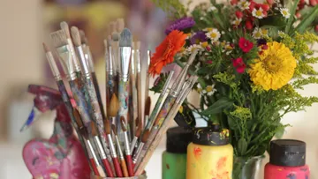 Brushes are placed in a container surrounded by flowers and paints.