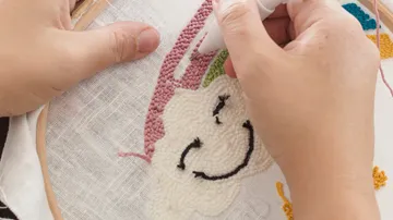 Hands embroider happy face on a fabric frame.