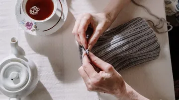 One hand knits a grey scarf with tea next to it.