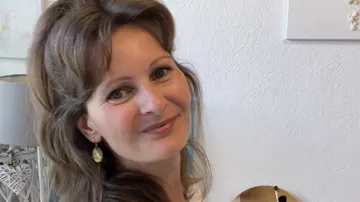Woman is smiling, standing in a bright, cozy room.