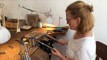 Woman works metal with a hammer at a table.
