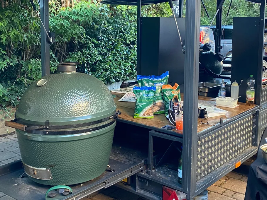 Green grill is on a cart surrounded by plants.