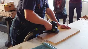 Ein Handwerker schleift Holz auf einem Tisch in einer Werkstatt.