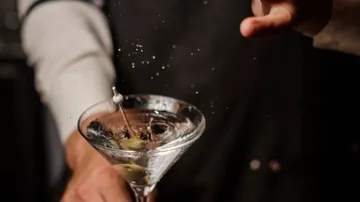 Martini-Glas mit Eis, Bartender gießt Cocktail mit Spritzern.