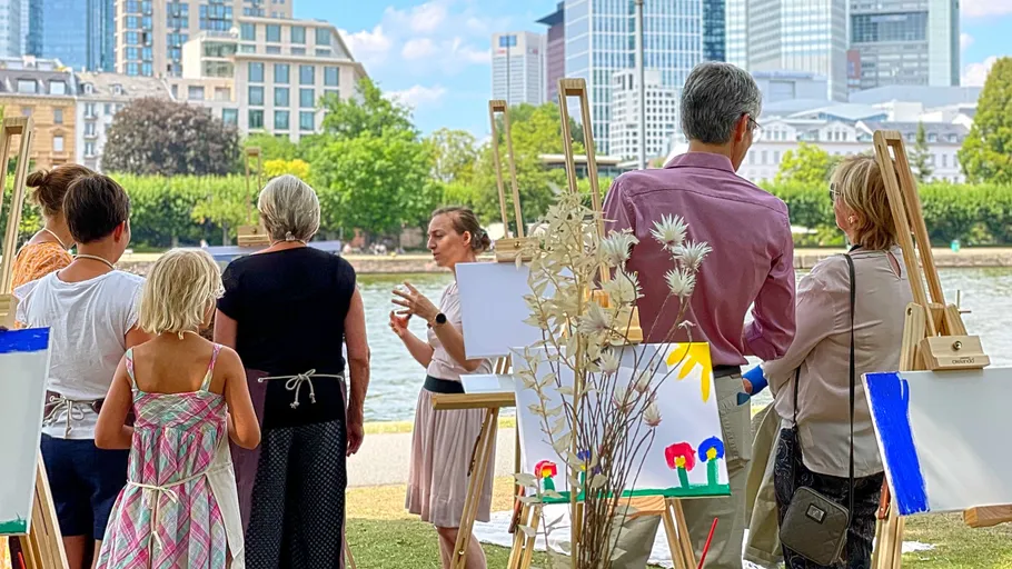 People paint on easels outside against a city backdrop on the water.