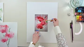 Kunstwerk: Hände malen rosa Blumen auf Papier. Malutensilien vorhanden.