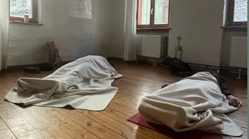 Zwei Personen liegen unter Decken auf Holzboden in einem Raum.