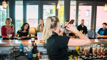 Barkeeperin schüttelt Cocktails, Gäste beobachten in modernem Barambiente.