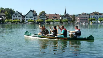 Green canoe on water, four people paddling, cityscape in the background.
