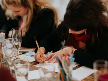 Two women draw with craft materials at a table.