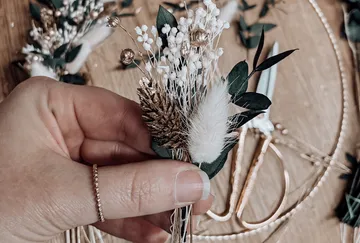Hand haltend ein kleines Blumenarrangement mit getrockneten Blumen und viel Grün, Schere und Bindfaden im Hintergrund.