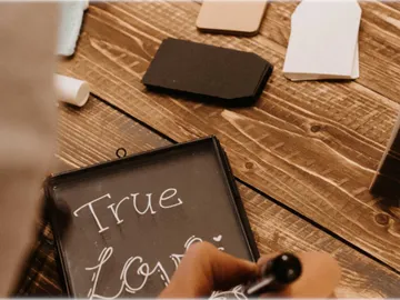 Eine Hand schreibt `True Love` auf eine Tafel, umgeben von Papier.