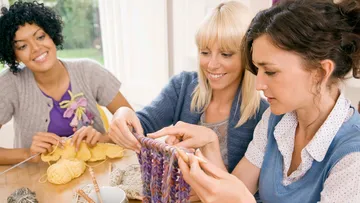 Drei Frauen stricken gemeinsam an einem Tisch in Innenräumen.