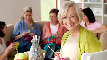 An elderly woman is happily knitting in a room with friends.