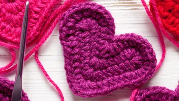 Purple crocheted heart is lying on white table.
