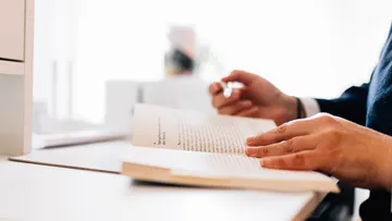 One hand holds a book and reads at a desk.