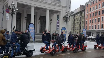 Gruppen von Menschen fahren auf roten Dreirädern vor einem Gebäude.