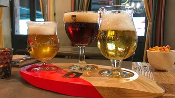 Three glasses of beer are standing on a wooden board in a nice environment.