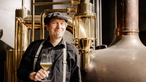 Mann mit Bierglas steht vor einer Brauanlage.