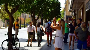 People walk on a sunny street, some take pictures.