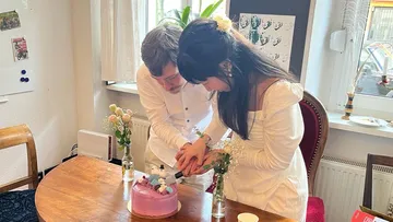 Two people cut a cake together in a room.