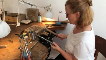 Woman works metal with a hammer at the table.