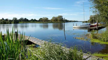 Holzsteg am ruhigen Wasser mit Booten im Hintergrund.