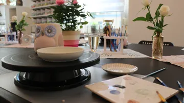 Ceramic figure sits on table surrounded by painting supplies and plants.