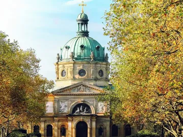 Kuppelgebäude steht zwischen bunten Laubbäumen unter blauem Himmel.