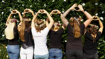 Six women shape hearts with their hands, surrounded by plants.