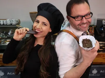 Two people smile, one eats chocolate, the other shows beans.