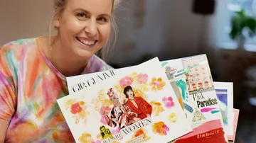 Smiling woman holding artistic prints in colorful surroundings.