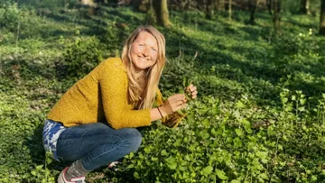 Woman collects herbs in a sunny, green forest.