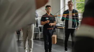 Wooden box hangs on colorful rubber bands, people watch.