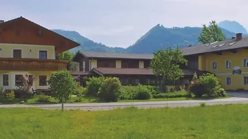 Bauernhäuser stehen in grüner Umgebung mit Bergen im Hintergrund.